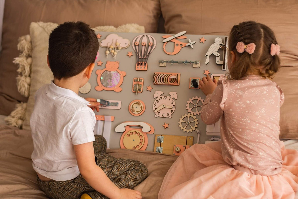 Deux jeunes enfants assis sur un lit qui manipulent une busy board en bois