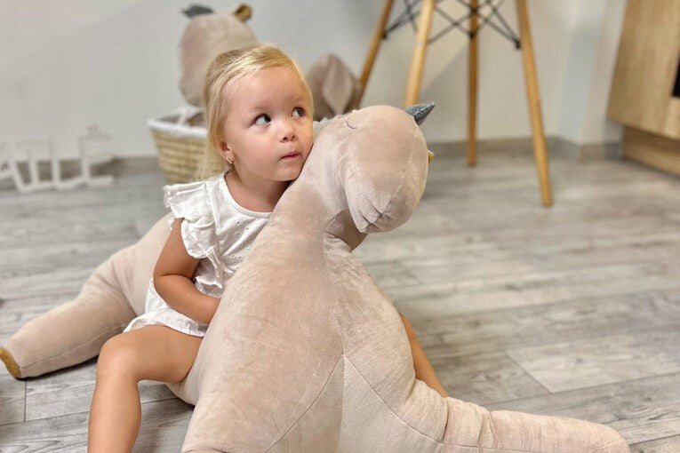 Fillette assise par terre qui enlace une grande peluche pégase en velours crème