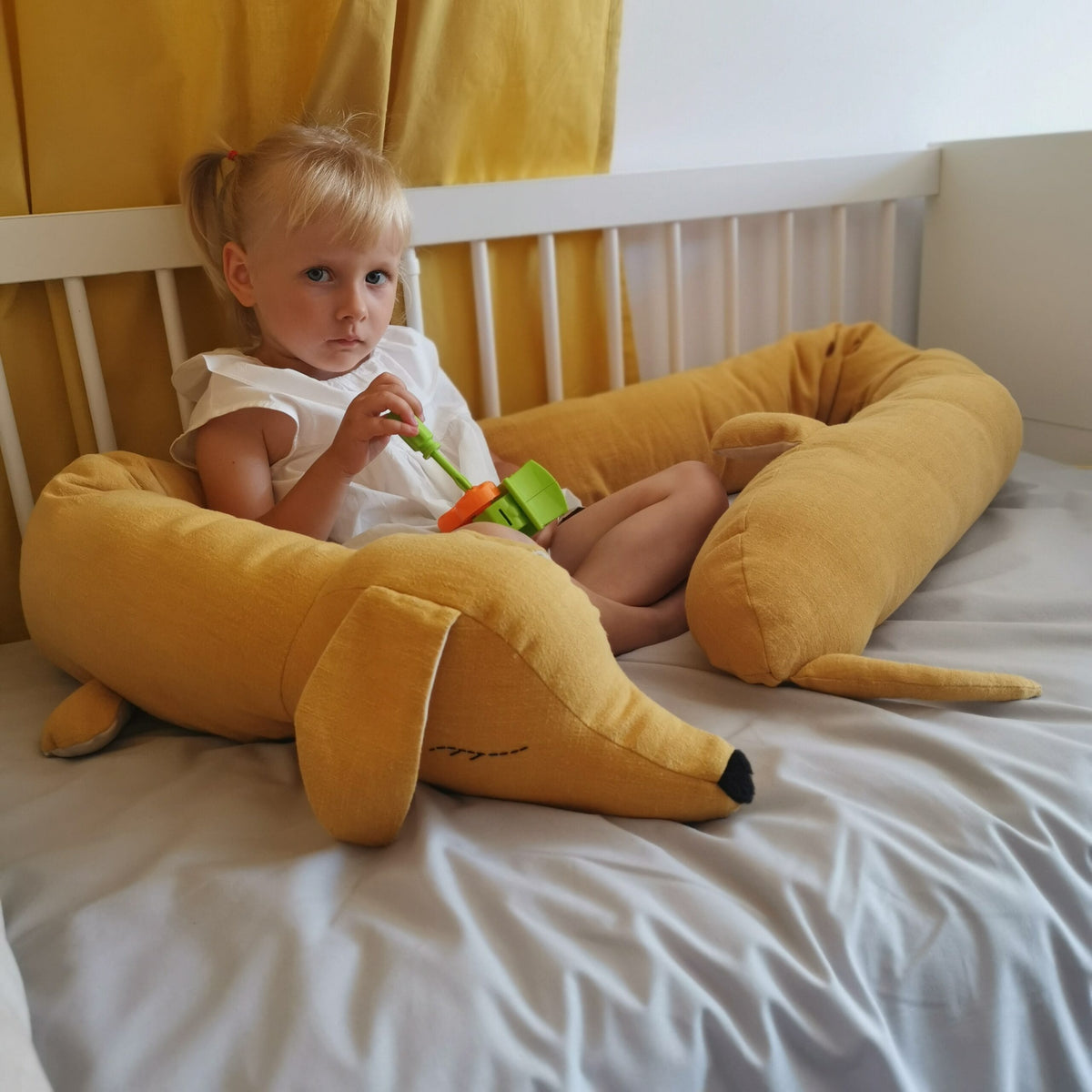 Une petite fille assise sur son lit avec un Dragonsaur CuddleRest moutarde