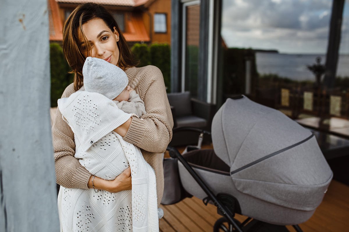 Une mère tient son bébé dans ses bras à côté d'une poussette Noordi grise